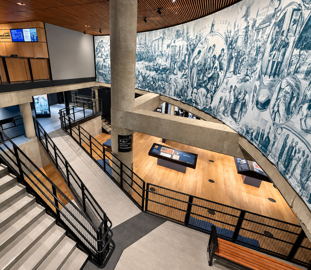 Looking down into a spacious, multilevel museum atrium built with exposed concrete, pale wood flooring, black metal railings, and a slatted wood ceiling. A broad staircase descends along our left edge beside an accessible ramp that zigzags toward the lower level. A thick cylindrical concrete column rises near the center, partly blocking the view. On our right, an enormous curved blue-and-white historical mural spans the upper wall and continues downward beside the open gallery. It contains densely arranged scenes of people, buildings, flags, boats, gatherings, and oval portrait-like vignettes. Below it, two low, dark display tables stand on a warm wood floor. A long wood-and-black-metal bench occupies the lower right foreground. At upper left, a wood-paneled counter or closed service area sits beneath orange and blue signs and digital screens. Track lights and recessed ceiling lights illuminate the otherwise empty interior.