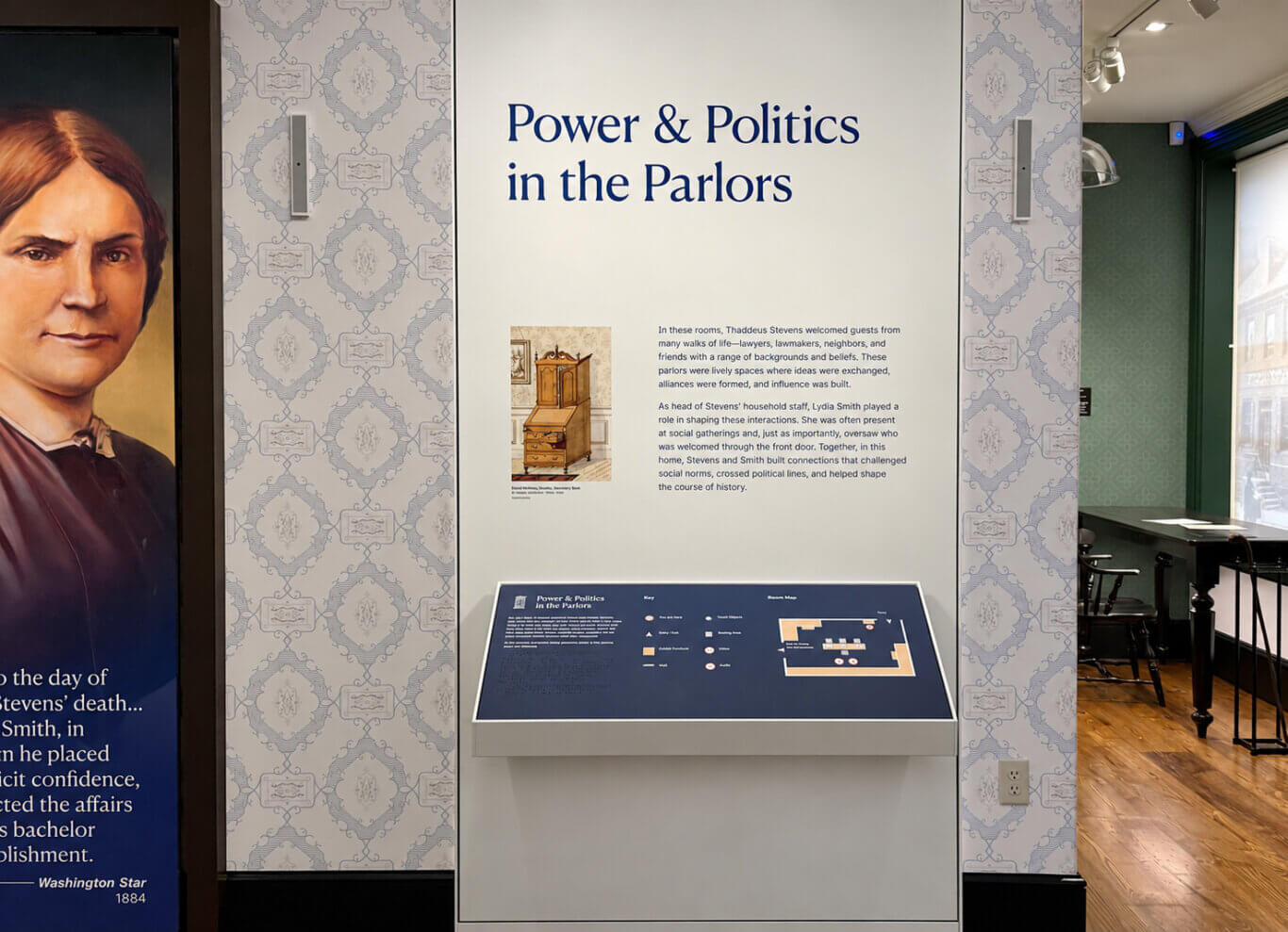 A museum exhibit stands against pale blue-and-white patterned wallpaper, centered on a large white wall panel above an angled interactive display and a portrait of Lydia Smith on our left. The main heading in large dark blue letters reads: "Power & Politics in the Parlors". Below it, an illustration of a wooden secretary desk appears beside two paragraphs that read: "In these rooms, Thaddeus Stevens welcomed guests from many walks of life-lawyers, lawmakers, neighbors, and friends with a range of backgrounds and beliefs. These parlors were lively spaces where ideas were exchanged, alliances were formed, and influence was built. As head of Stevens' household staff, Lydia Smith played a role in shaping these interactions. She was often present at social gatherings and, just as importantly, oversaw who was welcomed through the front door. Together, in this home, Stevens and Smith built connections that challenged social norms, crossed political lines, and helped shape the course of history." The dark blue angled display below repeats the heading "Power & Politics in the Parlors" and includes raised tactile text and Braille, a "Key," and a "Room Map." Legible key labels include "You are here," "Entry / Exit," "Exhibit Furniture," "Staff," "Touch Objects," "Seating Area," "Video," and "Audio"; the remaining small introductory text and map labels are too small to read. At the far left, a cropped painted portrait shows part of Lydia Smith, a mixed race woman with medium-light skin, reddish-brown hair, a dark garment, and a serious expression. Beneath the portrait, only fragments of a quotation remain visible. To the right of the exhibit, an adjoining room contains green patterned walls, a polished wooden floor, black chairs, and a tall dark table beside a large window. Two vertical wall lights flank the central panel, and ceiling spotlights illuminate the gallery.