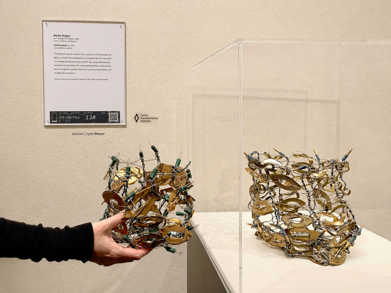 A museum display shows a wall label above and behind part of a sculptural bowl in a glass case made from tangled wire and found objects, with a person's hands holding a similar vessel in the foreground. The sculpture is an open, irregular vessel assembled from silvery wire, amber-gold curved fragments, small green bulb-like pieces, clear glass bulbs, and other small metal or plastic elements. A person wearing a black long sleeve reaches in from the lower left, with light-toned hands holding a very similar, but slightly smaller object. Above, the wall label is mounted in a clear frame with small dark fasteners at the corners. Its text reads: Marita Dingus, Born Seattle, Washington, 1966,, active in Auburn, Washington, Untitled Bowl, ca. 2005, wire and found objects, This delicate vessel, made of wire, strands of old Christmas tree lights, and other found objects, is a metaphor for the treatment of enslaved people of African descent. By using materials that are normally discarded, the artist celebrates the resilience and beauty of spirit in people who have had to overcome the harsh realities of colonialism., Boise Art Museum Permanent Collection, Gift of Ben and Aileen Kolin. At the bottom of the label, a black audio-access panel includes a phone icon, raised braille dots that are too small to read, the number 208-269-7051, the access code 13, and a QR code. To the right of the label, a diamond-shaped tactile icon appears beside text that reads: Tactile Representation Available.
