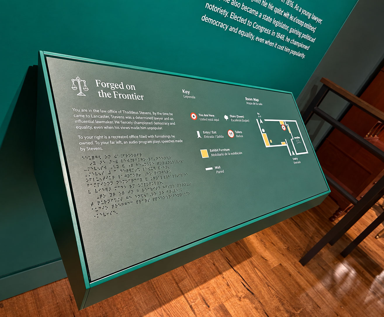 A dark green museum information panel and map titled Forged on the Frontier stands at an angle against a teal wall, with a polished wooden floor. A white scales-of-justice icon appears beside the title. On the left side of the panel, the main English text reads: "You are in the law office of Thaddeus Stevens. By the time he came to Lancaster, Stevens was a determined lawyer and an influential lawmaker. He fiercely championed democracy and equality, even when his views made him unpopular. To your right is a recreated office filled with furnishings he owned. To your far left, an audio program plays speeches made by Stevens." Several lines of raised Braille appear beneath this text. The key and map labels are in Spanish and English. The center heading reads "Key" and "Leyenda." Its bilingual labels read: "You Are Here" and "Usted está aquí"; "Stairs (Down)" and "Escaleras (abajo)"; "Entry / Exit" and "Entrada / Salida"; "Toilets" and "Baños"; "Exhibit Furniture" and "Mobiliario de la exhibición"; "Wall" and "Pared." Colored and white symbols accompany each label. On our right, the heading reads "Room Map" and "Mapa de la sala." The simplified floor plan uses white lines for walls, yellow blocks for exhibit furniture, and a red-and-white marker for the viewer's location. A north arrow is labeled "N," and the bottom of the map reads "Entry" and "Entrada." At the top of the wall, a paragraph is cropped by the image.