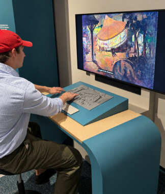 A seated museum visitor with light skin tone touches a slightly sloped grey tactile relief of a circus tent in front of a digital screen with an image of the painting on the screen. His fingertips rest on raised shapes and textured lines. The screen displays a colorful painting of a nighttime outdoor scene, with an orange-roofed carousel glowing among dark blue trees and small silhouetted figures; a caption at the bottom reads, "Couple in the Shadows." A black speaker is mounted directly below the screen. The visitor is wearing a red baseball cap, a pale blue-and-white striped shirt, and olive-green trousers.