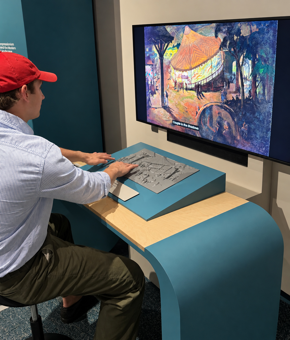 A seated museum visitor with light skin tone touches a slightly sloped grey tactile relief of a circus tent in front of a digital screen with an image of the painting on the screen. His fingertips rest on raised shapes and textured lines. The screen displays a colorful painting of a nighttime outdoor scene, with an orange-roofed carousel glowing among dark blue trees and small silhouetted figures; a caption at the bottom reads, "Couple in the Shadows." A black speaker is mounted directly below the screen. The visitor is wearing a red baseball cap, a pale blue-and-white striped shirt, and olive-green trousers.