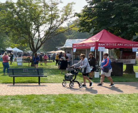 A sunny outdoor art market set up in a park. On our left, a white tent displays colorful paintings, including a large bear portrait and landscape-style artwork. Several people with light skin tone are walking on the grass in front of the booth. An additional cluster of people look at the art in the booth next to a parked wheelchair. In the background, there are more visitors, parked cars, a closed patio umbrella, and large leafy trees providing shade.