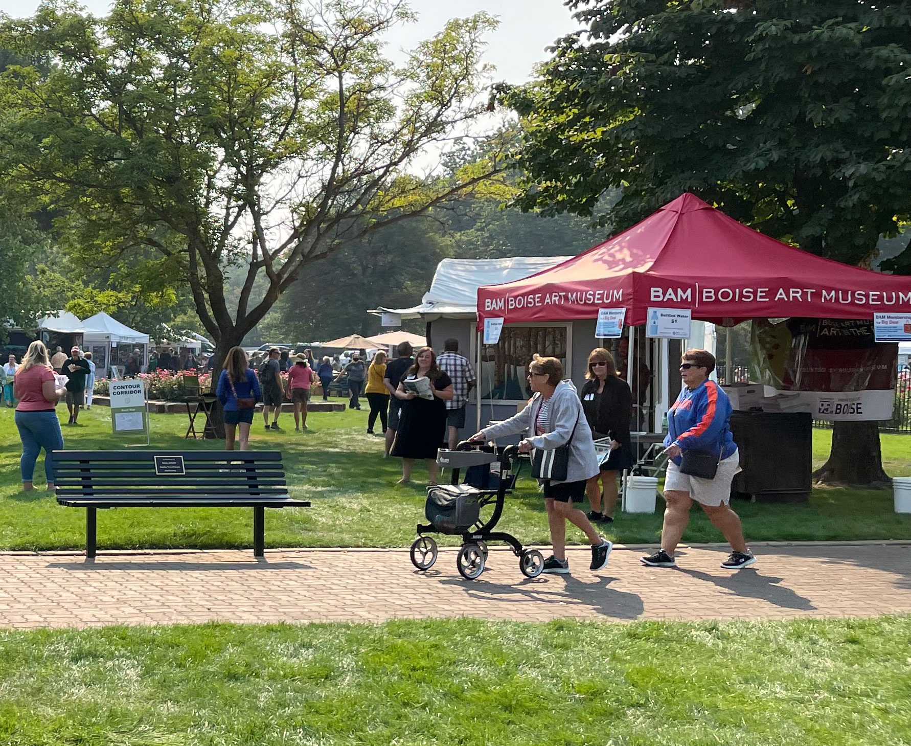 A sunny outdoor art market set up in a park. On our left, a white tent displays colorful paintings, including a large bear portrait and landscape-style artwork. Several people with light skin tone are walking on the grass in front of the booth. An additional cluster of people look at the art in the booth next to a parked wheelchair. In the background, there are more visitors, parked cars, a closed patio umbrella, and large leafy trees providing shade.