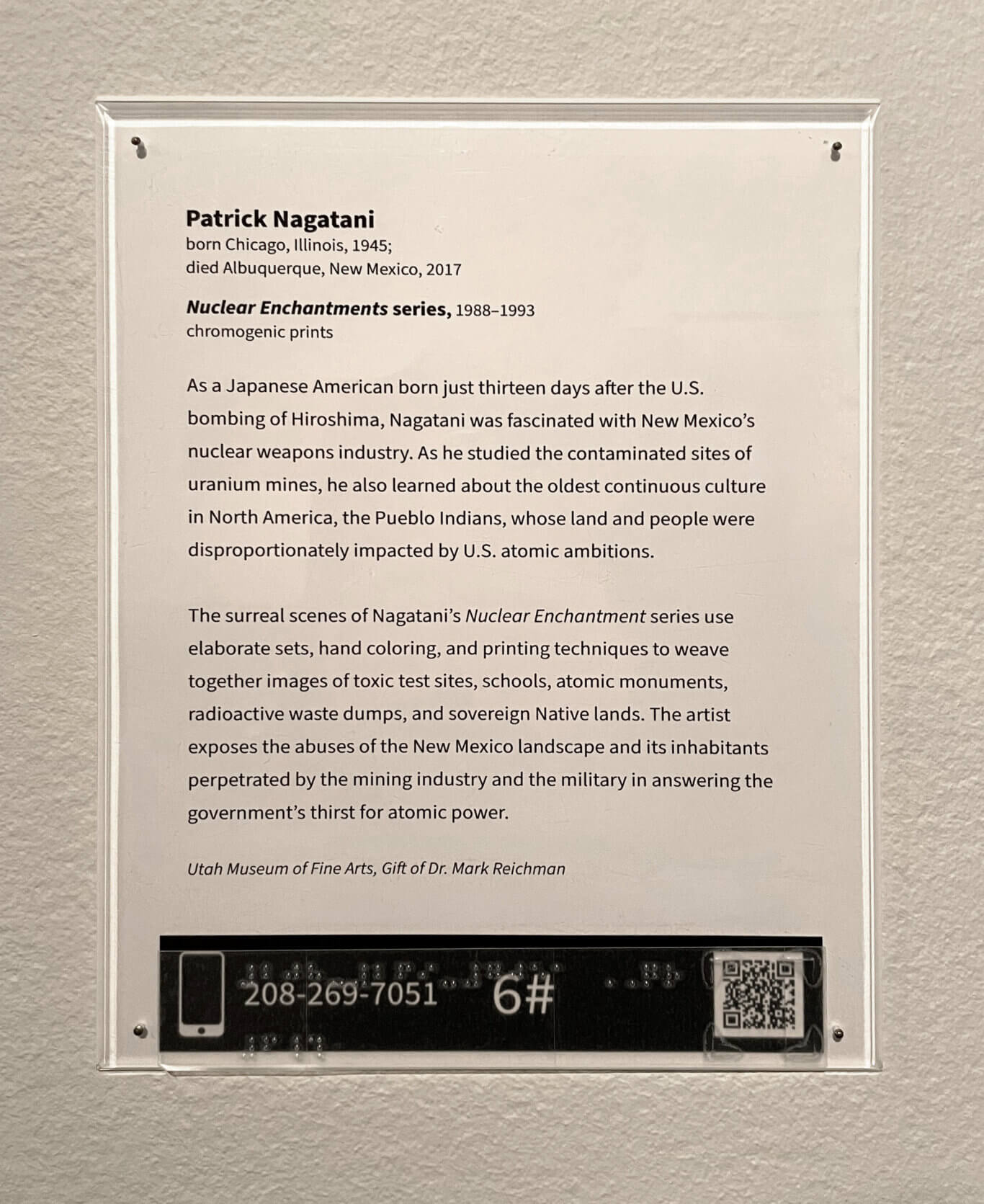 An acrylic-covered museum wall label for an artwork by Patrick Nagatani is mounted on a textured light gray wall; the white label has black printed text, and a black tactile/audio guide strip with raised Braille dots and a QR code along the bottom. The label text reads: Patrick Nagatani; born Chicago, Illinois, 1945; died Albuquerque, New Mexico, 2017; Nuclear Enchantments series, 1988-1993; chromogenic prints; As a Japanese American born just thirteen days after the U.S. bombing of Hiroshima, Nagatani was fascinated with New Mexico's nuclear weapons industry. As he studied the contaminated sites of uranium mines, he also learned about the oldest continuous culture in North America, the Pueblo Indians, whose land and people were disproportionately impacted by U.S. atomic ambitions. The surreal scenes of Nagatani's Nuclear Enchantment series use elaborate sets, hand coloring, and printing techniques to weave together images of toxic test sites, schools, atomic monuments, radioactive waste dumps, and sovereign Native lands. The artist exposes the abuses of the New Mexico landscape and its inhabitants perpetrated by the mining industry and the military in answering the government's thirst for atomic power. Utah Museum of Fine Arts, Gift of Dr. Mark Reichman. On the black strip at the bottom, visible printed text reads: 208-269-7051; 6#