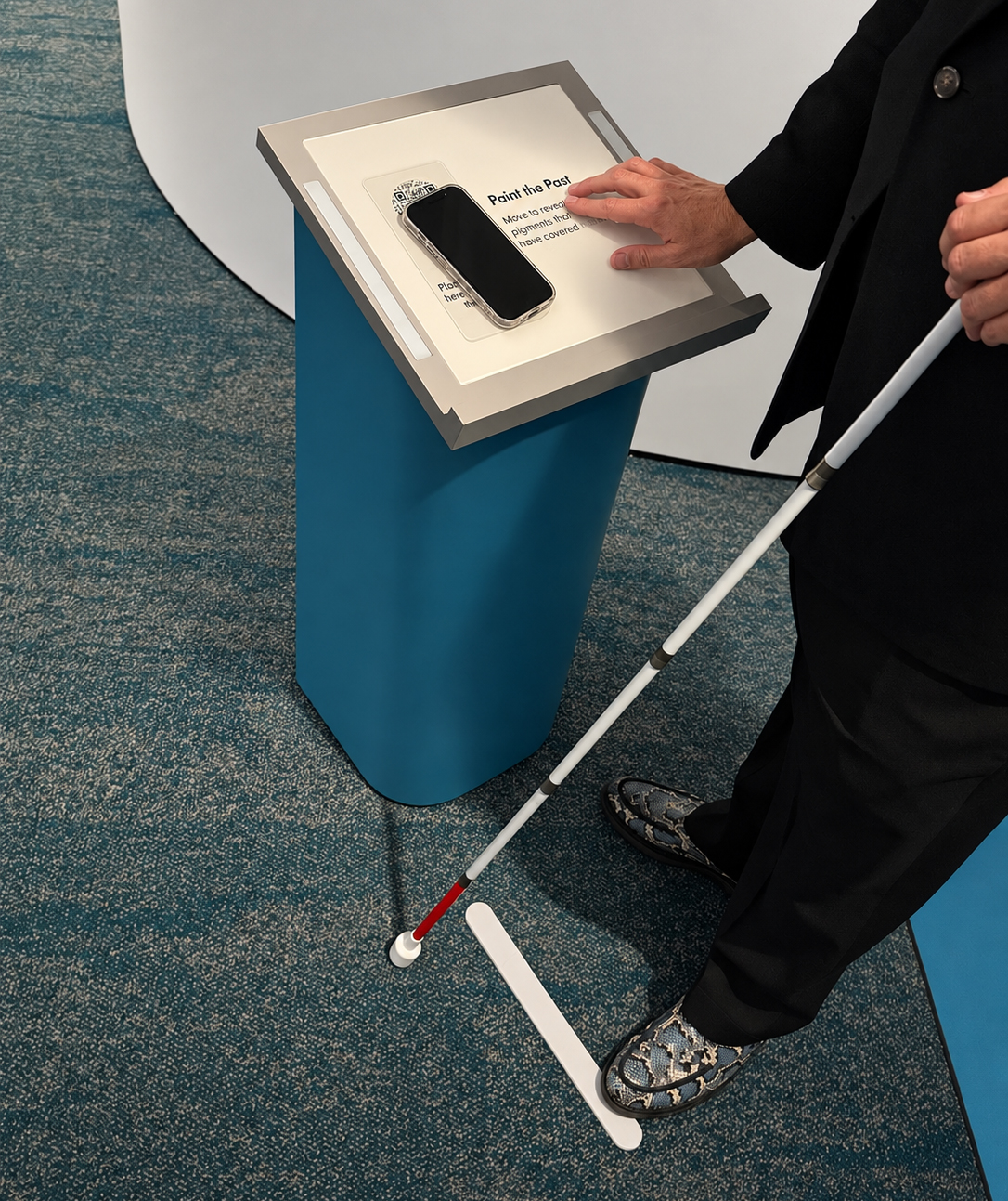 An adult with light skin reading Brailed on a short podium with a smart phone sitting on top while holding a white mobility cane. The person stands on our right in a black coat and trousers with gray, black, and blue snake skin patterned shoes. His left hand rests on the slanted rectangular display, which is mounted on a tall blue pedestal, while their right hand holds the cane. A black-screened smartphone in a clear case lies diagonally across the display, partly covering a QR code and printed instructions. The visible heading reads "Paint the Past." Beneath it, partly covered text reads "Move to reveal pigments that have covered"; the rest is obscured by the hand. Near the phone, another block of small text is largely obscured and unreadable. A long, raised white floor marker juts out from the podium and the person's shoe rests on the end. The floor has a mottled teal-and-beige carpet, and a curved white wall or display panel fills the background.