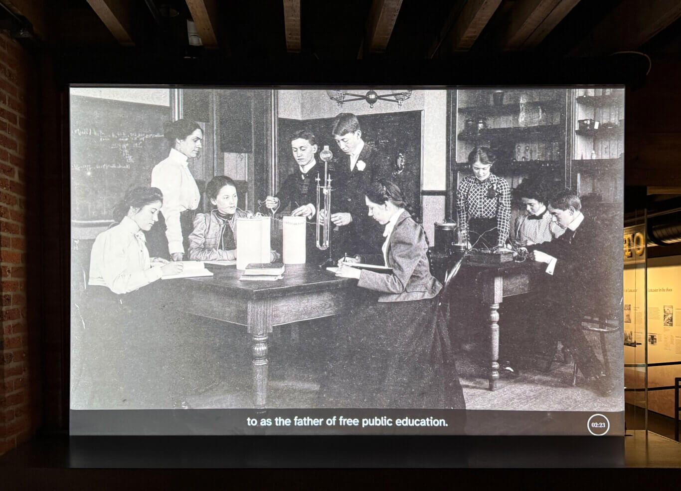 A large black-and-white historical photograph is projected across a screen in a dim brick museum interior. The photograph shows a crowded classroom with young adults with light skin tone seated closely together at long wooden desks, writing or reading, while several adults or older students stand among them. The room pictured has shelves, framed boards, tall windows, hanging lamps, and large suspended fixtures. Brick arches and dark ceiling beams remain visible above the projection, and a narrow opening through the screen reveals warmly lit museum displays behind it. A dark bar crosses the bottom of the projection, where the visible sentence fragment reads: "to as the father of free public education." At the right, a circular timer reads: "02:23".