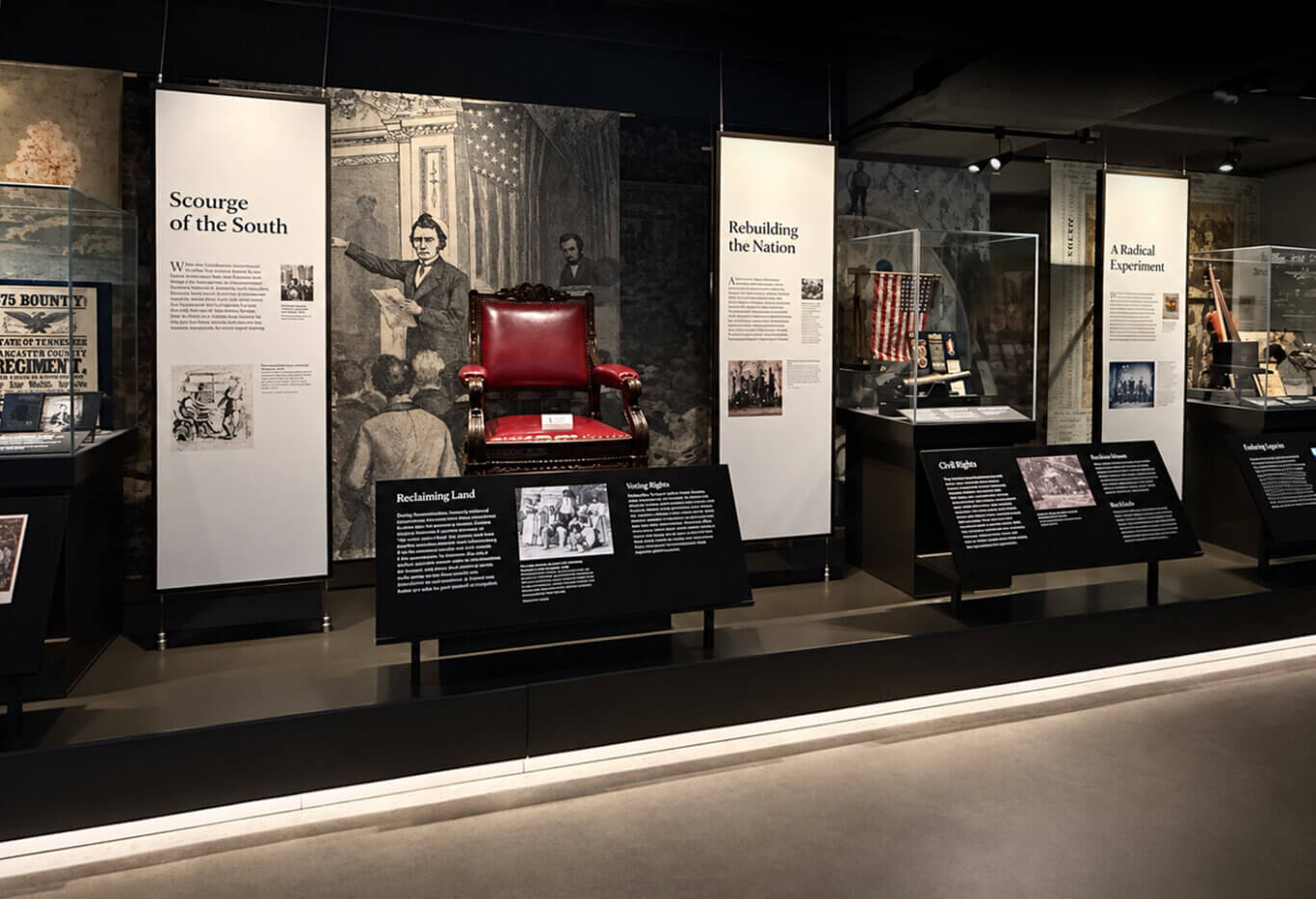 A dimly lit exhibit about the Reconstruction era, arranged on elevated black flooring with bright white floor lighting along the edge. Tall white interpretive panels, glass display cases, black angled text stands, historical images, and artifacts extend from our left to right beneath ceiling spotlights. Only the headings are large enough to read the text. At the center, an ornate dark wooden armchair upholstered in glossy deep red leather stands before a large monochrome mural of a formally dressed white speaker addressing a crowded chamber with an American flag behind him. The large panel to the left is headed "Scourge of the South." The black stand in front of the chair has the headings "Reclaiming Land" and "Voting Rights." A tall white panel just right of our center reads "Rebuilding the Nation." Beside it, a glass case contains a vertically draped red, white, and blue American flag, framed items, and several small objects on angled supports. The black stand in front of this case is headed "Civil Rights." Farther right, another tall white panel reads "A Radical Experiment." Additional glass cases hold long wooden objects, documents, photographs, and other artifacts. At our far right, a cropped black stand is headed "Enforcing Justice." Dark walls covered with enlarged historical illustrations form the background, while a pale gray walkway fills the foreground.