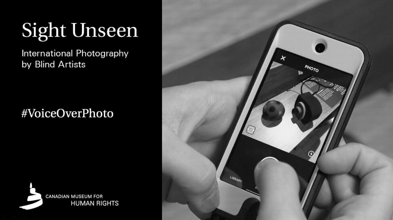 A black-and-white promotional graphic is divided vertically between a solid black text panel on our left and a close-up photograph of a smart phone on our right. Large white text at the upper left reads: "Sight Unseen". Beneath it, smaller text reads: "International Photography by Blind Artists". Midway down the panel is the hashtag "VoiceOverPhoto". At the bottom, a white abstract logo resembling a curled ribbon or candle appears beside the words "CANADIAN MUSEUM FOR HUMAN RIGHTS". On our right, two light toned hands hold a smartphone at an angle while a thumb touches its screen. The phone displays a camera interface photographing a tabletop with a small spotted ball, a dark angular device with a circular center, coiled cables, and a printed page whose tiny text is too blurred to read. Legible interface text includes an "×" at upper left, "PHOTO" near the top, "LIBRARY" at lower left, and a partially obscured "P" near the bottom center. Circular interface icons appear near the lower corners of the displayed photograph.