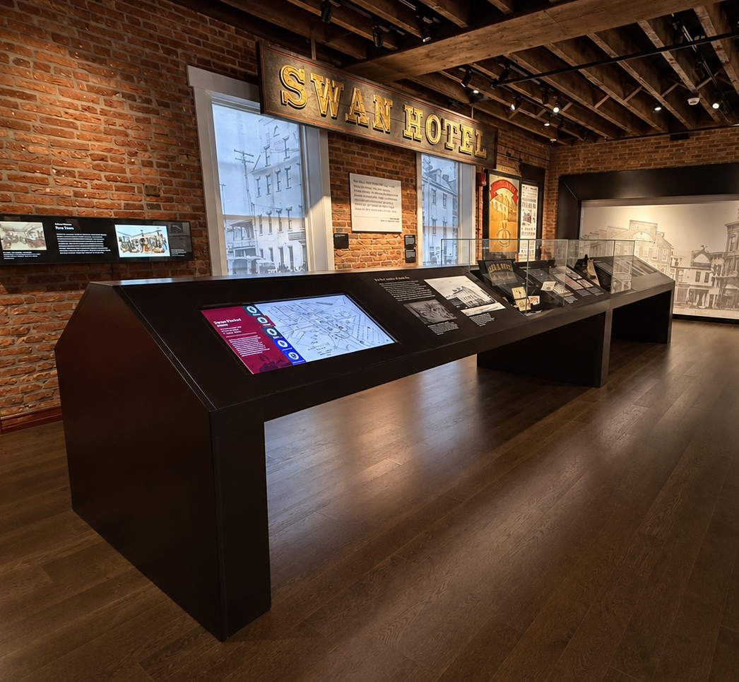 A wide-angle photograph shows a museum gallery, with exposed red-brick walls, dark wood flooring, open ceiling beams, and black track lighting. A large weathered sign above two tall windows reads "SWAN HOTEL" in illuminated gold letters. Long, angular black exhibit tables extend from the foreground toward the back wall; the nearest includes a touchscreen map with a red information panel, while farther displays contain photographs, documents, small artifacts, and angled interpretive labels beneath glass. A horizontal digital display is mounted on the left brick wall, and a framed red-and-yellow poster hangs between the windows and the rear exhibit. At right, a large pale mural depicts a historic streetscape and hotel building. Snowy or overcast building exteriors are visible through the windows.