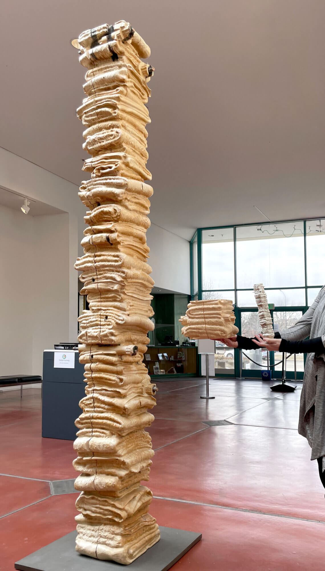 A tall pale wood sculpture stands on a dark rectangular base in a spacious museum lobby, with a rough vertical column made of many carved, layered, folded-looking forms resembling blankets. The main column fills the left-center of the image and extends beyond the top of the frame; its surface is cream and golden brown, with chisel marks, natural grain, cracks, seams, knots, and some darkened end areas. To our right, a partially cropped adult, visible from the torso and hands only, holds two related carved wooden pieces: a blocky horizontal section with the same dimensions as the original sculpture on one palm and a thinner vertical stacked piece in the other hand. Behind them are red-brown floors, white walls, high ceilings, gallery lights, green-framed windows and doors, and another similar carved column or segment farther back.