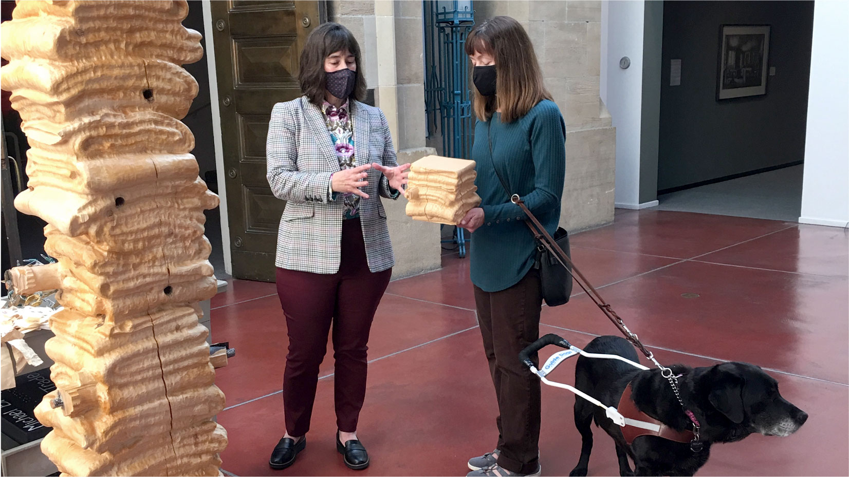 A gallery scene shows two masked adults with light skin tone standing beside a large pale carved wooden form while one holds a smaller matching carved block, with a black guide dog in a harness on our right. On our left, the tall wood sculpture of stacked blankets rises out of the frame and has stacked, wavy, horizontal ridges, visible cracks, knots, and protruding pegs or rods; its warm blond surface catches bright light. Near the center, Melanie Fales, a white woman with shoulder-length dark hair gestures with both hands toward the smaller block as if she's about to grab it. On our right, another woman with straight shoulder-length brown hair holds the small carved block at waist height while a black crossbody bag hangs at her side. The black dog stands in the lower right facing right, wearing a brown and white guide harness; the visible blue text on the harness reads, "Guide Dogs." The setting has a reddish polished floor, pale stone architectural walls, a dark wooden door, a blue metal gate, and an open doorway into a darker gallery where a framed picture hangs on the wall.