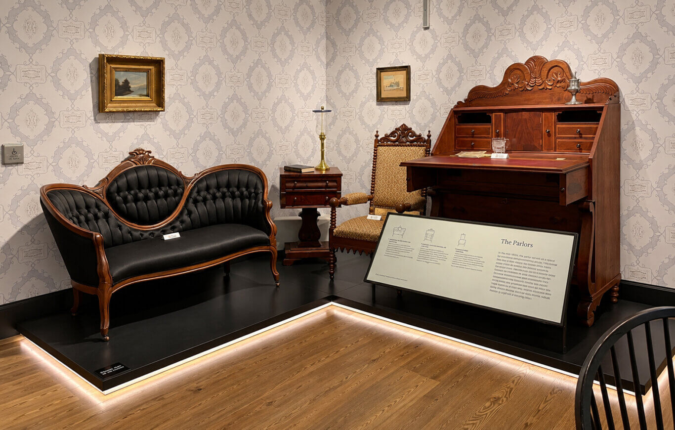 A museum display recreates a Victorian-style parlor with ornate dark wood furniture arranged on a raised black platform against pale gray patterned wallpaper. On the left stands a carved wooden settee upholstered in deeply tufted black leather, its back formed by three rounded sections. A small white object label rests on the seat, but its text is too small to read. Above it hangs a gold-framed landscape painting of trees beside water. Near the center, a two-drawer wooden side table supports several stacked books and a tall brass oil lamp with a clear glass chimney. Behind and to the right is a carved armchair with patterned gold-brown upholstery and a small white label whose text is illegible. A smaller framed picture hangs above the chair. Dominating the right side is a large reddish-brown wooden secretary desk with an elaborate carved crest, multiple small drawers and compartments, a lowered writing surface, and larger drawers below. Papers, a faceted glass vessel, a small white label, and a silver-colored decorative object sit on or above the desk. In front of the furniture, a large angled interpretive panel displays three small line drawings of chairs and a prominent title reading "The Parlors"; all additional body text is illegible. Warm strip lighting outlines the platform above the honey-colored wood floor.