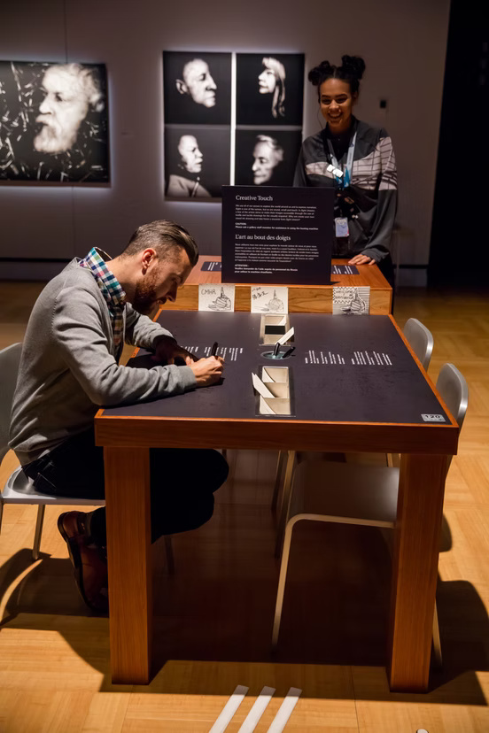 A color photograph shows two adults at an interactive museum table in a softly lit portrait gallery. In the foreground at left, Rob Itri-Vincent, a Metí man with light skin, short brown hair, and a beard leans over the black tabletop, using both hands to write on a card. Behind the table laughing is a standing person with medium-brown skin and dark hair arranged in two high buns wearing a lanyard. The large wooden table has a black display surface printed with blocks of small white text, most of which are too blurred to read, and several raised or recessed geometric elements aligned down its center. A black upright panel at the far end is headed "Creative Touch." Paper samples attached to the wooden edge include the legible letters "CMR"; other printed and handwritten markings are indistinct. Gray chairs surround the table, and three white tactile floor strips approach it from the bottom edge. On the wall behind the visitors hang black-and-white portrait photographs: at our left, a large close-up profile of an older person with white hair and a full white beard; at center, four smaller profile portraits arranged in a two-by-two grid, with pairs of faces turned toward one another.