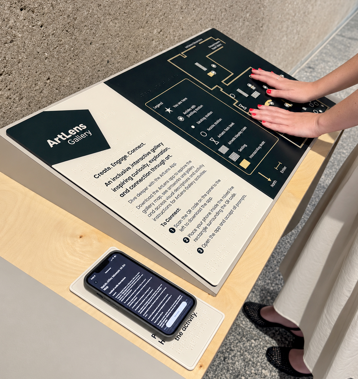An ArtLens Gallery tactile map table viewed from above and at an angle, with a large black-and-cream tactile map, raised Braille, and a smartphone resting on a smaller panel. At right, a person with light skin tone, red-painted fingernails, and a silver ring places both hands on the map. The large panel's dark logo block reads "ArtLens" and "Gallery." Beneath it, bold text reads: "Create. Engage. Connect. An inclusive, interactive gallery inspiring curiosity, exploration, and connection through art." Smaller instructions read: "Dive deeper with the ArtLens App. Download the ArtLens app to explore the gallery map, see artworks up close, and easily access audio descriptions and activity instructions for ArtLens Gallery activities." A section headed "To connect:" continues: "1 Scan the QR code on the panel to the left to download the app. 2 Place your phone inside the iridescent rectangle surrounding the QR code. 3 Open the app and accept all prompts." The black map uses thin gold outlines for rooms and white, gold, and gray accessibility symbols. Its key reads "Legend," "You are here," "ArtLens Exhibition Digital Display," "Drawing station," "Practice station," "Manual door button," "Seating," and "Interactive art"; one additional legend label and the small room names at the top are too blurred to transcribe reliably. Direction labels at the lower right read "North" and "Entrance." Numerous lines of raised Braille accompany the printed information, but the individual Braille characters cannot be reliably transcribed at this resolution. The smartphone shows a dark webpage whose heading appears to read "ArtLens Mobile Museum of Art." The table has a pale wood surface beside a rough gray wall and speckled gray floor.