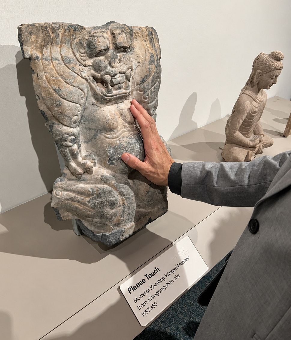 A museum display features a weathered gray-beige stone model of a kneeling winged monster, which a person touches with an open hand. The blocklike sculpture stands upright on a low beige platform against an off-white wall. Its high-relief figure has a broad head, bulging eyes, a wide toothy grin, two downward-pointing tusks, curling decorative forms, large wings framing its upper body, a rounded belly, bent legs, and worn, mottled surfaces. At right, only part of the visitor is visible: a light-toned hand rests flat against the monster's chest and abdomen, extending from a gray suit sleeve with a black cuff. Farther back on the right sits a smaller pale stone figure of a seated Buddha with wavy hair gathered in a bun, layered robes, a bowed head, and hands resting on his lap. An angled white label at the front reads in Braille and text: "Please Touch" followed by "Model of Kneeling Winged Monster" and "from Xianglongshan site" and "1957.360".