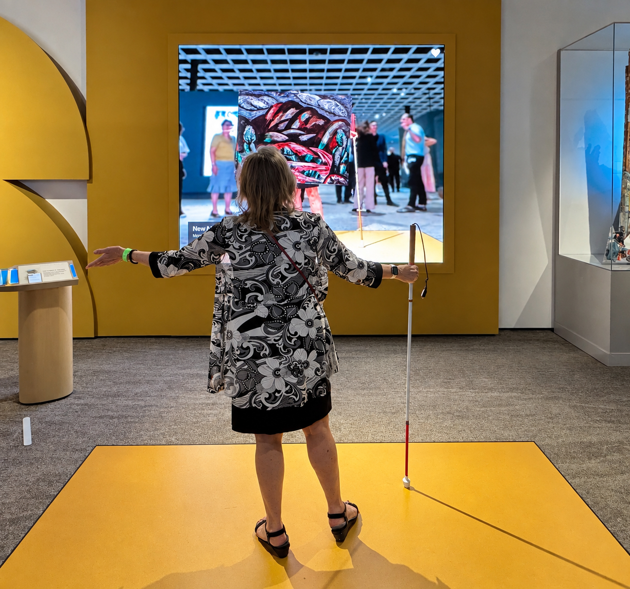 An adult from behind, standing on a large yellow platform in a museum gallery and facing a wall-mounted interactive screen. The visitor has light skin and shoulder-length light brown hair and wears a loose black, white, and gray floral-patterned top over a black skirt or dress. Their arms extend outward to either side; their right hand holds a long white cane with a red end, its tip resting on the platform. The screen displays a blurred gallery scene with several visitors beneath a white grid ceiling, overlaid at center by a vivid rectangular artwork filled with twisting red, black, white, turquoise, and blue forms. A small white heart icon appears in the screen's upper-right corner. The display is set into a mustard-yellow wall framed by large curved yellow architectural panels on the left. A small pedestal on our far left holds several labels or controls with blue-lit edges. On our right, a tall glass display case glows blue and contains only a cropped portion of an object near the image edge. Gray patterned carpet surrounds the yellow flooring.