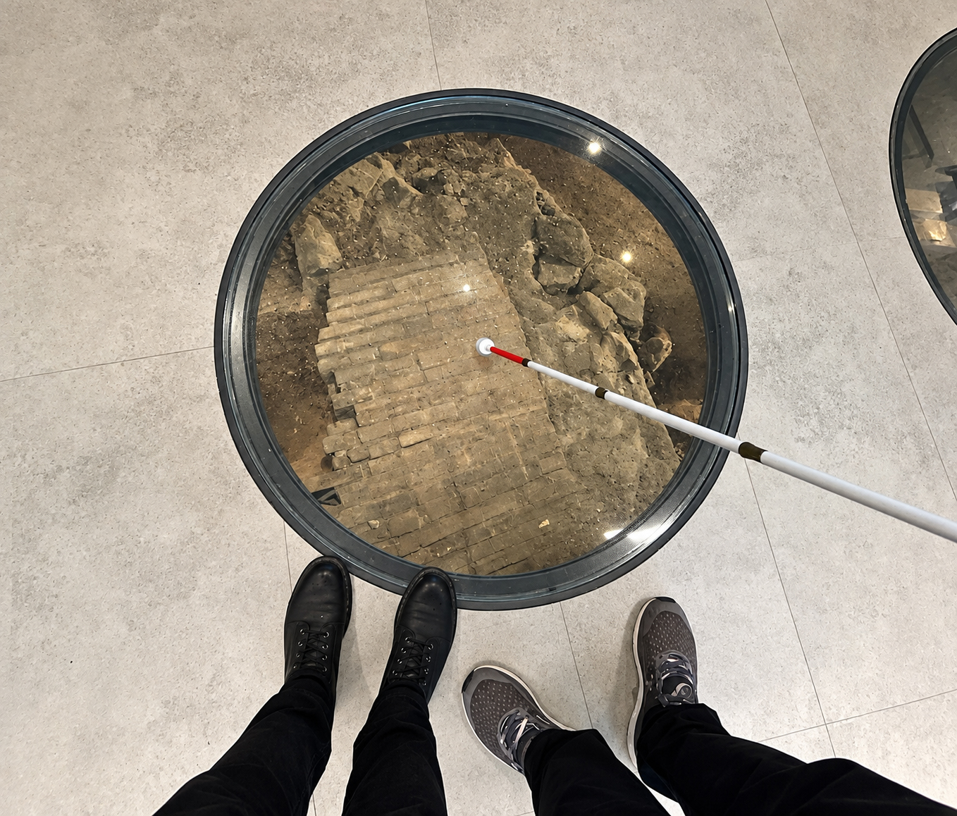 A top-down photograph shows a large circular glass panel set into a pale gray tiled floor, revealing an archaeological excavation below. Beneath the glass are packed brown earth, irregular stones, and a rectangular section of old beige brickwork laid in uneven rows. Reflections of several ceiling lights dot the glass. A white cane extends diagonally from our lower right, with its white tip touching the glass near the center. At the bottom edge, two people stand beside the panel: one wears black pants and black lace-up boots, while the other wears black pants and gray lace-up athletic shoes. A small portion of another circular glass panel appears at our upper right.
